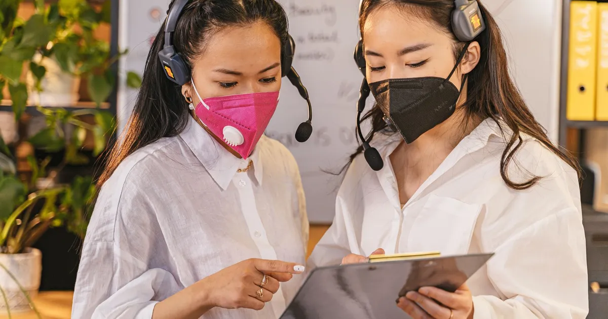 Two women in face masks and headsets discussing work on a clipboard in an office setting.