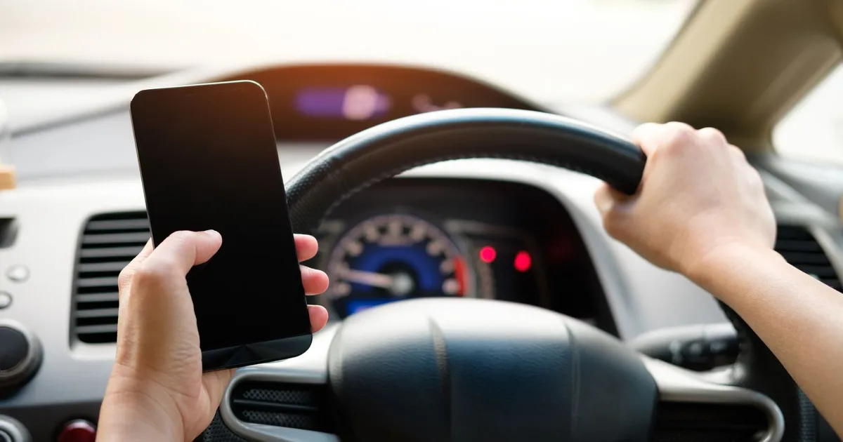 A driver uses a smartphone while steering a vehicle, showcasing distracted driving risks.
