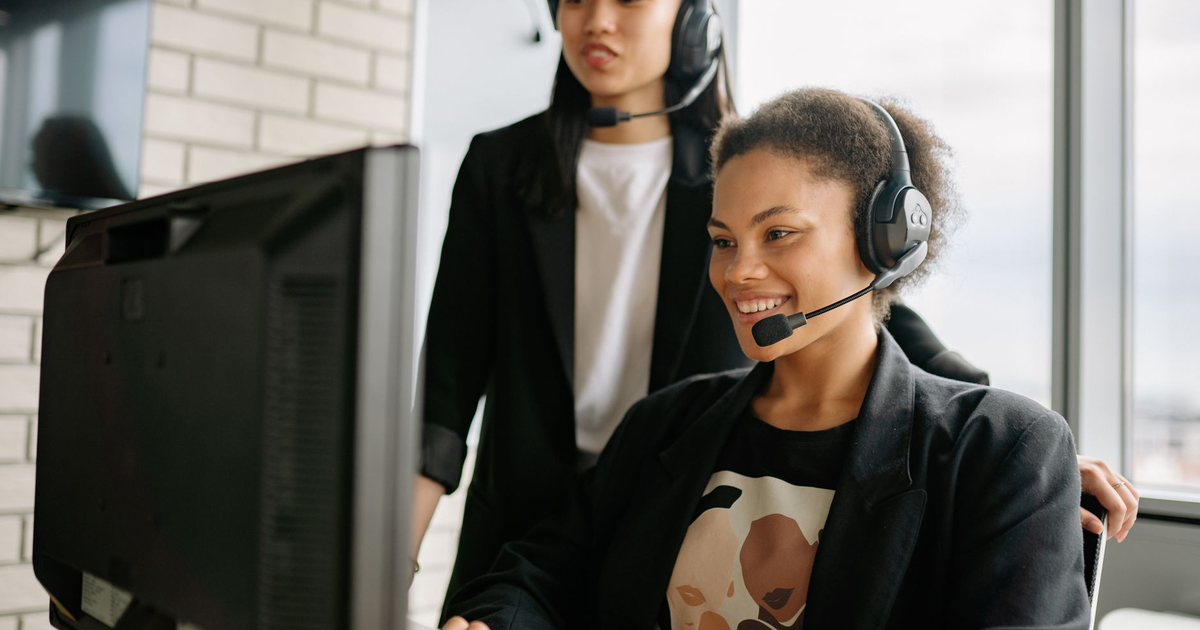 Two women with headsets engaged in customer service at a computer in a bright office.