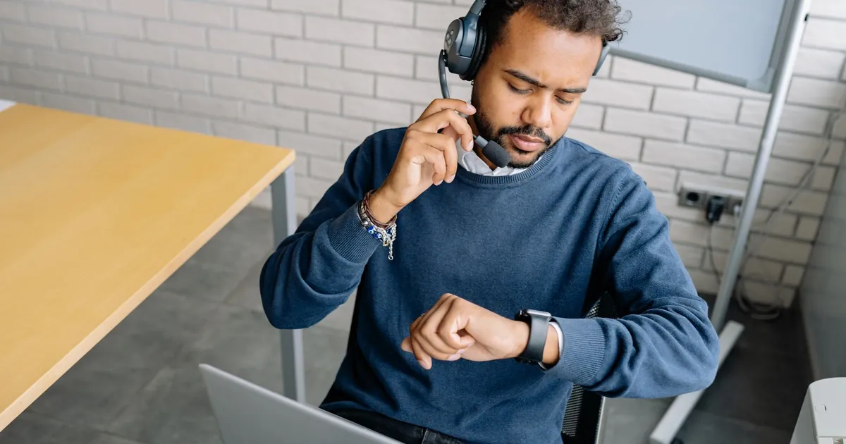 A focused call center agent wearing a headset checks the time on his wristwatch in a modern office setting.